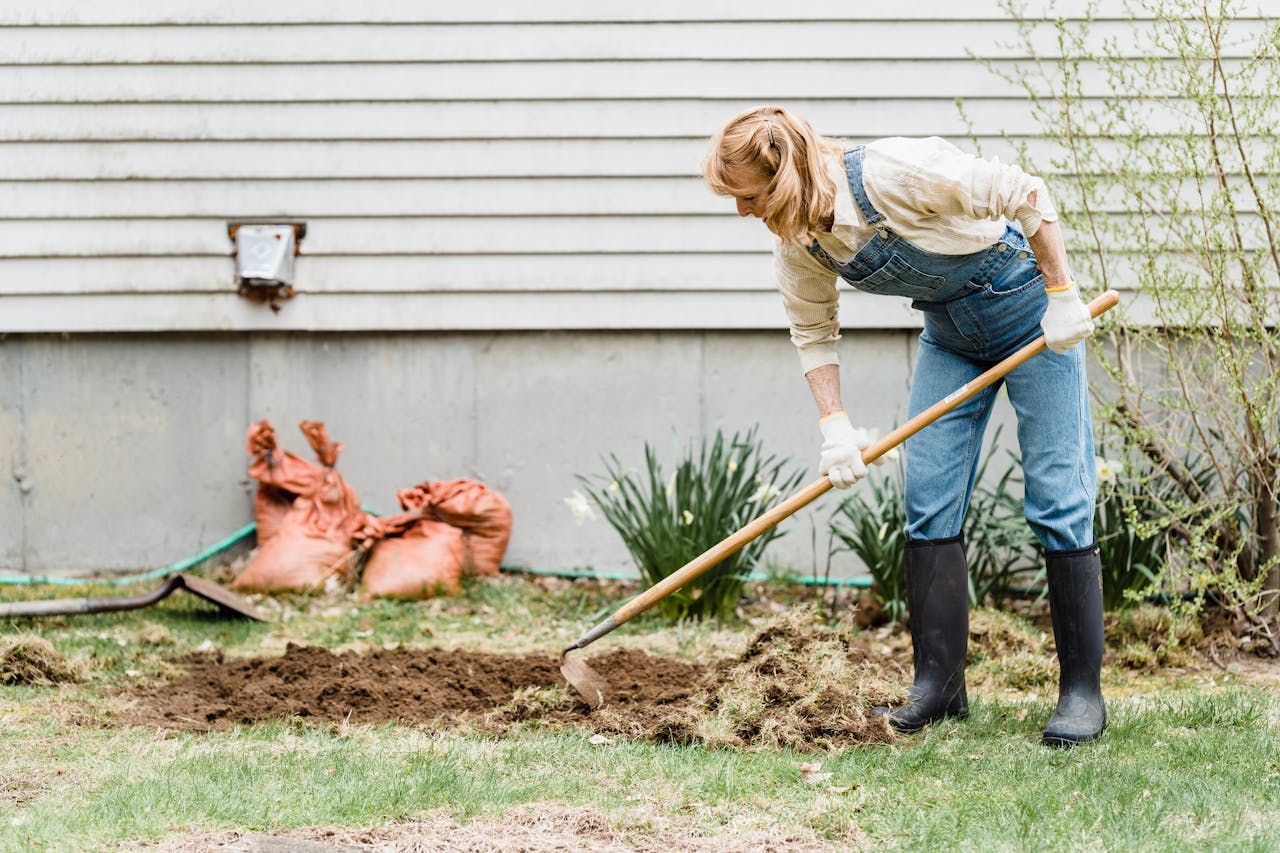 Woman in denim overalls working in the garden, tending soil with a hoe.